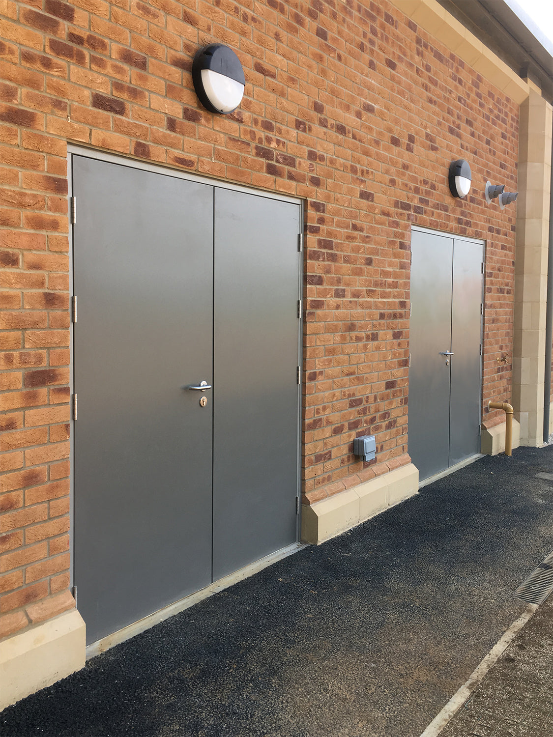 Two gray fire rated doors on a brick building with a dark asphalt driveway.
